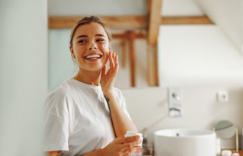 Beautiful young woman taking care of skin by applying moisturizer cream in bathroom