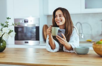 A woman in a cozy kitchen enjoys her morning coffee while browsing her smartphone. Fresh flowers and fruits create a serene and inviting atmosphere, perfect for a relaxing start to the day.