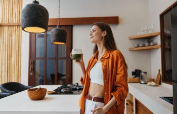 Young woman enjoying a green smoothie in a modern kitchen, wearing an orange shirt and white pants, radiating happiness and wellness in a bright, stylish setting