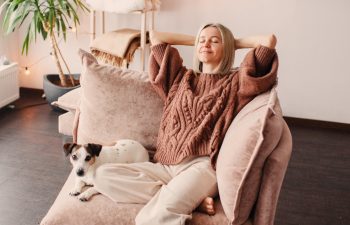 A woman sits relaxed on a sofa with her hands behind her head, eyes closed, while a small dog lies beside her.